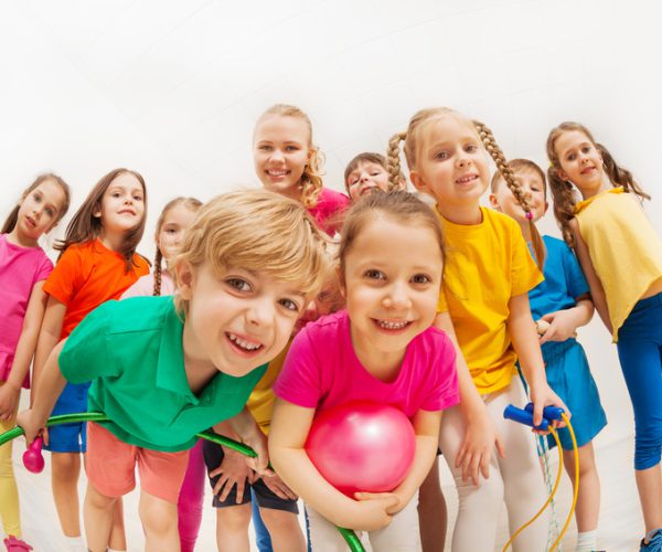 Portrait of cute kids in sportswear with female gymnastics coach, holding sports equipment, standing together in gym, looking at camera